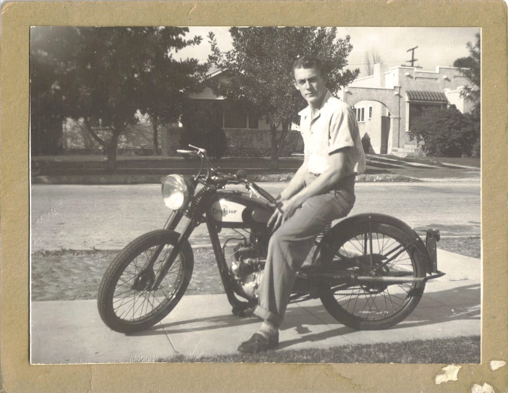 My grandfather as a young man, seated on his Excelsior motorcycle on a sunlit street, 1940s.